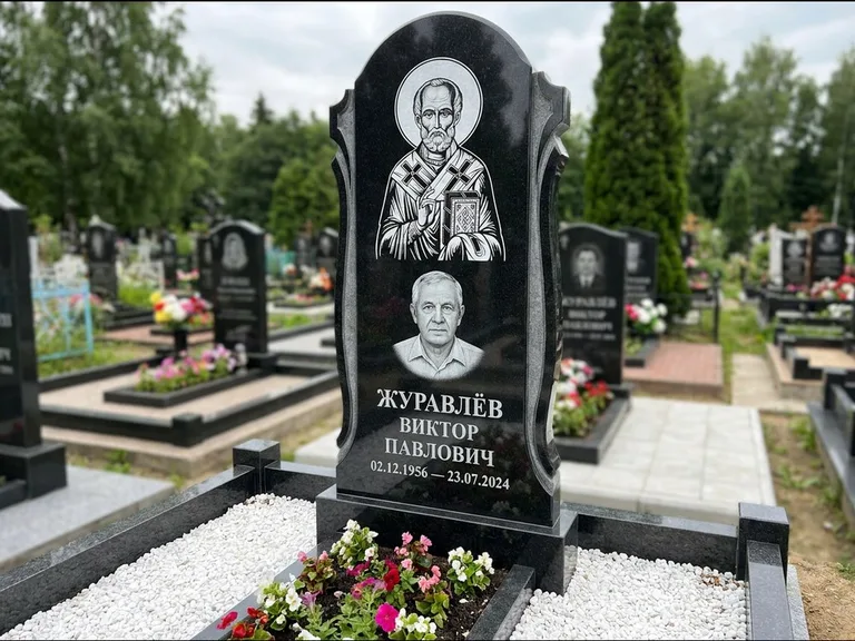 Памятник с гравировкой иконы: стела с образом в полный рост на обороте — гравировка образа Матроны Московской, низкие кусты спиреи в цвету, работа «Лебедева» в Томске